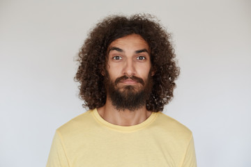 Fototapeta na wymiar Studio shot of pretty brown-eyed dark haired curly male with beard wearing yellow t-shirt while standing against white background, looking to camera with calm face
