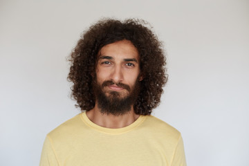 Fototapeta na wymiar Indoor photo of pleasant looking positive young bearded man with brown curly hair smiling slightly while posing over white background, wearing casual clothes