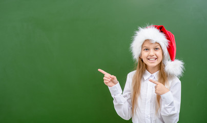 Smiling girl wearing a red christmas hat points away on empty chalkboard. Space for text