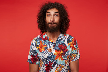 Fototapeta na wymiar Surprised attractive brunette curly guy with beard biting his lips and looking to camera with raised eyebrows, standing over red background with hands along body