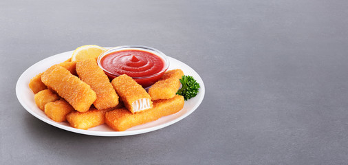 Fish fingers sticks on a plate with parsley, lemon and ketchup on a gray texture.