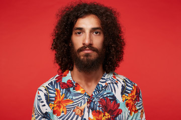 Portrait of handsome brown-eyed young bearded man with dark curly hair looking to camera with calm face and keeping his lips folded, isolated over red background in casual clothes