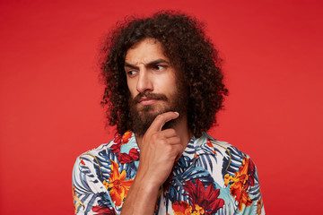 Fototapeta na wymiar Studio photo of pensive young brunette curly male with lush beard looking thoughtfully aside and frowning eyebrows, keeping raised hand on his chin while posing against red background