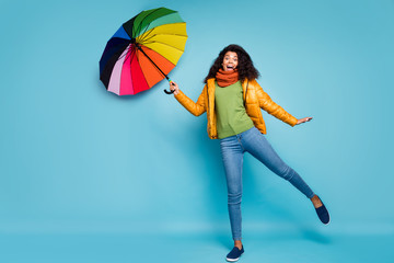 Full body photo of funny dark skin wavy lady hold parasol windy weather walk street rejoicing wear yellow coat jeans green sweater knitted scarf isolated blue color background © deagreez