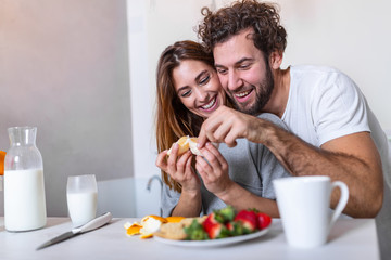 Obraz na płótnie Canvas Beautiful young couple is looking at each other and feeding each other with smiles while cooking in kitchen at home. Loving young couple embracing and cooking together, having fun in the kitchen