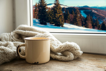 Winter background - cup with candy cane, woolen scarf and gloves on windowsill and winter scene outdoors. Still life with concept of spending winter time at cozy home with cold weather outdoors