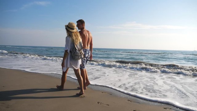 Couple of lovers walking at coast holding hands during resting on resort. Man and woman strolling on beach and enjoying summer vacation together. Honeymoon of young pair. Concept of love and happiness