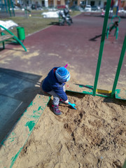 A little boy in a blue jacket and a hat with a pompom sits on the edge of the sandbox and plays...