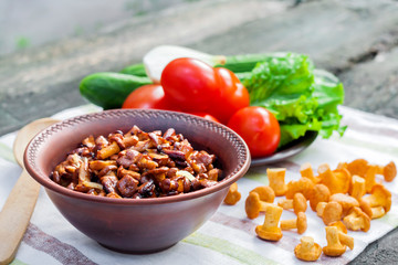 Fried chanterelles with onion in rustic bowl and plate with fresh vegetables for salad on background