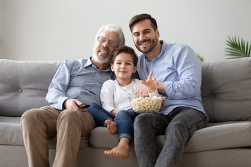 Cheerful grandfather grownup son and grandchild watching TV eating popcorn