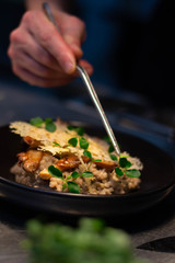 a head chef cooks buckwheat and wild mushroom risotto, served with herbs, white wine and cheese on top. The chef uses a tweezers to decorate the dish 