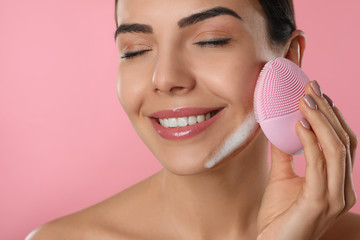 Young woman using facial cleansing brush on pink background, closeup. Washing accessory