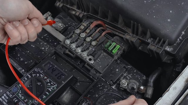Man measures DC voltage on terminals of automotive battery using multimeter in winter daytime in parking. Malfunction or problem with automobile. Hood or bonnet raised up. Closeup of lead-acid battery