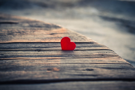 Lonely red heart on a table of old wood. Small red heart in the rays of the setting sun on an old wooden surface. Tender picture with a red heart on the beach. A little red heart lit by the setting su