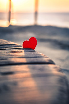 Small red heart in the rays of the setting sun on an old wooden surface. Tender picture with a red heart on the beach. A little red heart lit by the setting sun casts a shadow on the old wood.