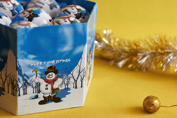 Box with Christmas tree toys, balls with a snowman on a yellow background.