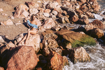 A girl with a luxurious figure poses on a rocky seashore in a cute blue swimsuit. Happy girl on vacation.