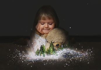 Little girl with her teddy bear, watching a enchanted forest  on the pages of an open magic book