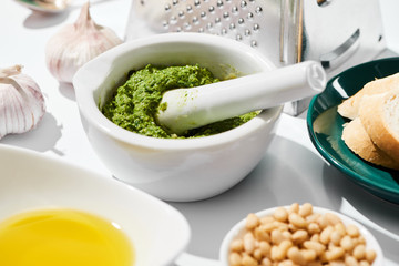 close up view of pesto sauce in pounder near grater, bread and ingredients on white background