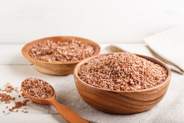 Two wooden bowls with unpolished brown rice and wooden spoon on a white wooden background. Side view, close up.