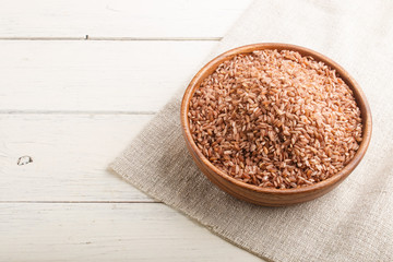 Wooden bowl with unpolished brown rice on a white wooden background. Side view, copy space.