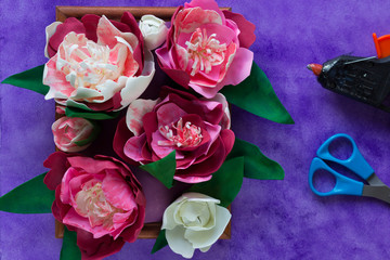 Handmade decorative peonies made from foamiran on wooden panel and glue gun and scissors on purple background