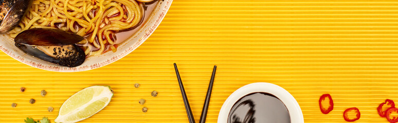 top view of seafood ramen near fresh soy sauce and chopsticks on yellow surface, panoramic shot