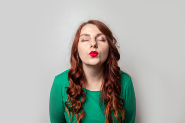 Young girl with red curly hair and red lipstick giving a kiss on a gray wall background