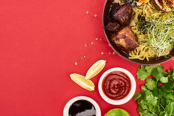 top view of spicy meat ramen near sauces, parsley and lime isolated on red