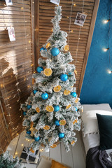 snow-covered Christmas tree decorated with blue balls and dried oranges. wood in the interior with screen, light floor and blue wall
