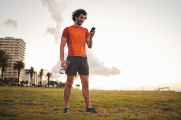 Full body shot of a Happy athletic young man holding water bottle in hand listening music on earphones using smart phone while standing in the park