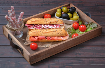 Serving or kitchen wooden tray with traditional Spanish ham sandwiches with slices fuet and jamon next to olives and ripe cherry tomatoes. Typical breakfast, tapas, snacks and appetizers.