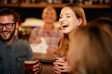 Gorgeous caucasian woman laughing and holding pint of beer while standing at pub surrounded by her best friends.