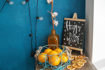 whole and cut oranges on the table, a vase with a twig and a Board with the inscription happy new year on the nightstand against the blue wall with a garland