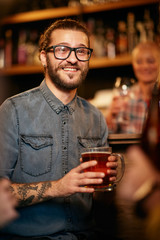 Handsome smiling hipster man sitting at pub near bar counter, holding pint of beer and listening interesting stories. Nightlife.