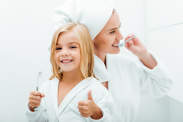 cute child holding toothbrush and showing thumb up near mother