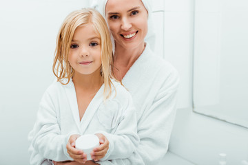 happy kid holding container with cosmetic cream near cheerful mother in bathrobe