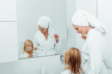 Happy mother looking at daughter brushing teeth near mirror