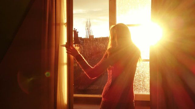 girl opening a window and looking at the evening city at the beams of orange sunset sun