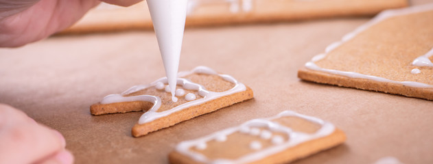 Woman is decorating gingerbread cookies house with white frosting icing cream topping on wooden table background, baking paper in kitchen, close up, macro. - obrazy, fototapety, plakaty