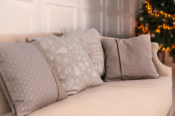 Luxury Christmas interior elements: beige cushions with Christmas pattern on beige old style couch in white room on decorated Christmas tree background