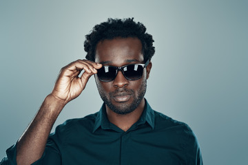 Handsome young African man looking at camera and adjusting eyewear while standing against grey background