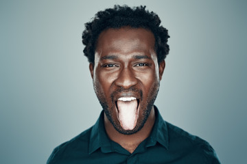 Handsome young African man looking at camera and sticking out tongue while standing against grey background