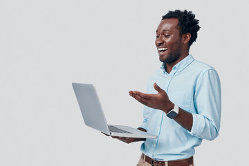 Handsome young African man using laptop and smiling while standing against grey background