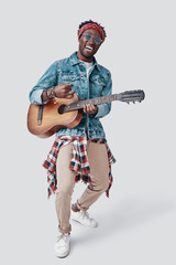 Full length of handsome young African man playing guitar and smiling while standing against grey background