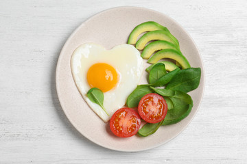 Plate of tasty breakfast with heart shaped fried egg on white wooden table, top view
