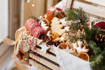 Girl is holding big wooden box with hand made christmas family presents: gingerbread cookies, christmas decor, knitted napkin. Cozy home atmosphere, holiday mood, christmas lights on. Closeup