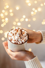 Young girl in knitted white wool sweater is holding a mug with hot chocolate or coffee with marshmallow. Christmas lights on, cozy holiday atmosphere, aromatherapy for cold winter season. Closeup
