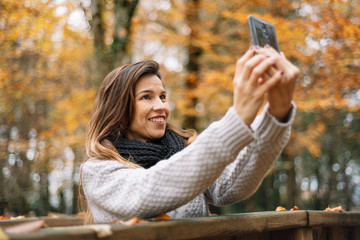 Beautiful young happy woman taking selfie with smartphone in autumn park. Season, technology and people concept .