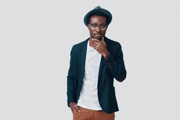 Handsome young African man looking at camera and keeping hand on chin while standing against grey background